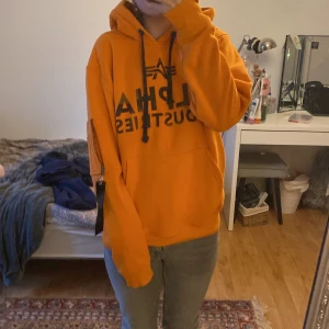 Orange hoodie från Alpha Industries - Säljer en cool orange hoodie från Alpha Industries med svart tryck på framsidan. Den har en stor luva och dragsko samt en rymlig passform. Perfekt för en avslappnad stil.