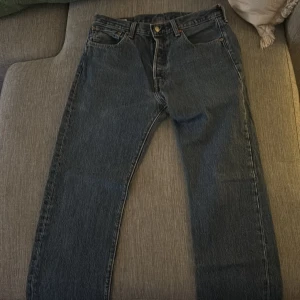 Levi's 501 - Säljer ett par klassiska blå jeans från Levi's med fem fickor och knappgylf. De har en rak passform och är perfekta för en avslappnad stil. Levi's-loggan syns tydligt på baksidan.