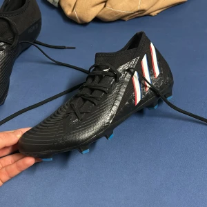 Svarta fotbollsskor från Adidas - Snygga svarta fotbollsskor från Adidas med snörning och tre ränder i vitt, rött och blått på sidan. Skorna har en mönstrad sula för bra grepp på planen.