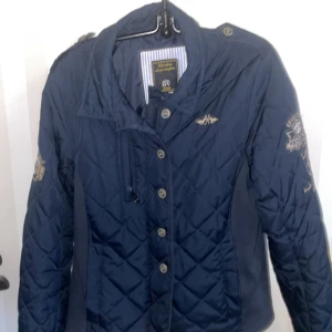 Mörkblå quiltad ridjacka från HV Society - Snygg mörkblå quiltad ridjacka från HV Society med broderade detaljer och knappar. Jackan har en sportig look med emblem och nummer på ärmen. Perfekt för en stilren och avslappnad stil.