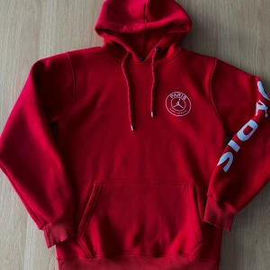 Röd hoodie från Paris Saint-Germain - Snygg röd hoodie med stor Paris Saint-Germain-logga på ryggen och ärmen. Hoodien har en klassisk känguruficka och justerbar huva med dragsko. Perfekt för sportiga tillfällen eller avslappnad stil.