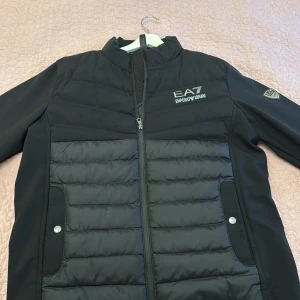 Armani jacka - Snygg svart jacka från Emporio Armani med dragkedja och quiltad design. Jackan har långa ärmar och två fickor med knappar framtill. Perfekt för kyligare dagar.