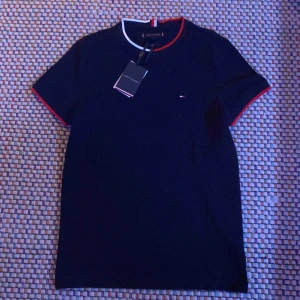 Svart t-shirt från Tommy Hilfiger  - Snygg svart t-shirt från Tommy Hilfiger med röda och vita detaljer vid krage och ärmslut. Den har år i passformen *slimfit* och är perfekt för en stilren look👑. Den är i storlek XS, men passar mig bättre som en S. Hör av dig✌️