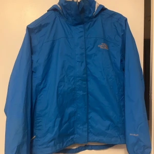 Blå vindjacka från The North Face - Säljer en blå vindjacka från The North Face med huva och dragkedja. Perfekt för blåsiga dagar och utomhusaktiviteter. Jackan har en enkel design med The North Face-logga på bröstet.