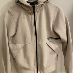 Beige hoodie från Sail Racing - Säljer en stilren beige hoodie från Sail Racing med dragkedja och huva. Jackan har en diskret logga på bröstet och en större på huvan. Perfekt för en avslappnad stil.