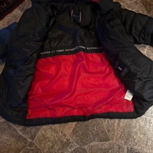 Svart dunjacka från Tommy Hilfiger - Säljer en svart dunjacka från Tommy Hilfiger med röd insida. Jackan har en klassisk pufferdesign och är perfekt för kalla dagar. Den har långa ärmar och en bekväm passform. Märkeslogga synlig på insidan.