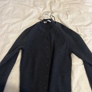 Mörkgrå ribbad kofta från Zara - Säljer en stilren mörkgrå kofta från Zara i ribbat material. Koftan har långa ärmar och knappar framtill, perfekt för lager-på-lager under kyligare dagar. Ett mångsidigt plagg för garderoben!