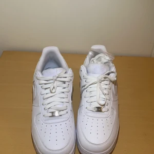 Vita Nike Air Force 1 nocta - Säljer ett par klassiska vita Nike Air Force 1 med snörning och stilren design. Skorna har en präglad Nike-logga på hälen och texten 'Love you forever' på sulan. Perfekta för en clean look.