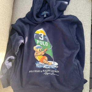 Mörkblå hoodie från Ralph Lauren - Säljer en mörkblå hoodie från Ralph Lauren med en cool Polo Bear-design som surfar. Tröjan har en klassisk passform med huva och ribbade muddar. Perfekt för en avslappnad stil.