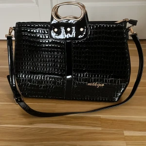 Svart axelväska från Makgio - Snygg svart axelväska i krokodilpräglat syntetmaterial från Makgio. Väskan har en elegant design med guldfärgade detaljer och en praktisk dragkedja. Insidan är rymlig med flera fack för enkel organisering. 