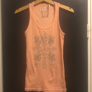 Persikofärgad linnetopp från Guess Jeans - Snygg persikofärgad linnetopp från Guess Jeans med glittrande stenar i ett dekorativt mönster på framsidan. Perfekt för att lägga till lite bling i garderoben. Toppen har en rund halsringning och ärmlös design.