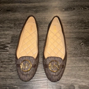 Bruna loafers från Michael Kors - Snygga bruna loafers från Michael Kors med ett quiltat mönster och en stor MK-logga framtill. Skorna har en klassisk design med rundad tå och är perfekta för en stilren look.