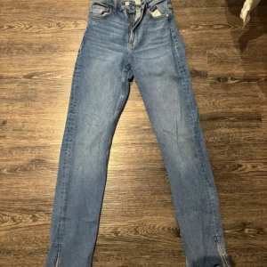 Blå jeans från Bershka - Snygga blå jeans från Bershka med hög midja och slitsdetaljer vid anklarna. Perfekta för en trendig look. De har en klassisk femficksdesign och är tillverkade i denim.