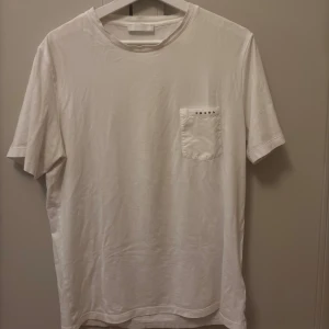 Prada T-shirt - Säljer en stilren vit t-shirt från Prada med en bröstficka. Perfekt för en minimalistisk look. T-shirten har en klassisk passform och är kortärmad. Ingenting og, bra condition.                                          Storlek L, men passar verkligen som en M.