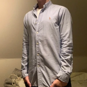 Blå skjorta från Ralph Lauren - Snygg blå skjorta från Ralph Lauren med broderad logga på bröstet. Skjortan har en slim passform och är tillverkad i bomull. Perfekt för en stilren look med knappar hela vägen ner och klassisk krage.