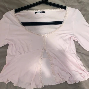 Ljusrosa topp från Gina Tricot - Söt ljusrosa topp från Gina Tricot med spetsdetaljer och en liten rosett framtill. Toppen har trekvartsärm och en fin, feminin design. Perfekt för en romantisk look.