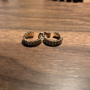 Guldiga örhängen med glittriga stenar - Snygga guldiga örhängen i creol-stil med glittriga stenar runt om. Perfekta för att ge lite extra bling till din outfit. De är små och eleganta, vilket gör dem lätta att bära hela dagen.