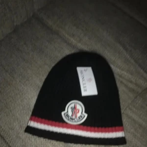 Moncler mössa !! - Snygg svart mössa från Moncler med röd och vit randdetalj. Perfekt för kyliga dagar med stilren design och Moncler-logga framtill.