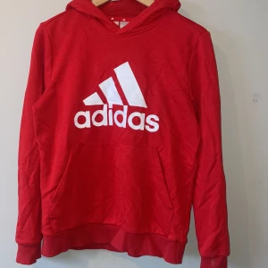 Röd hoodie från Adidas - Säljer en snygg röd hoodie från Adidas med det klassiska logotyptrycket i vitt på framsidan. Tröjan har en bekväm huva och en praktisk magficka. Perfekt för en sportig look eller avslappnade dagar.