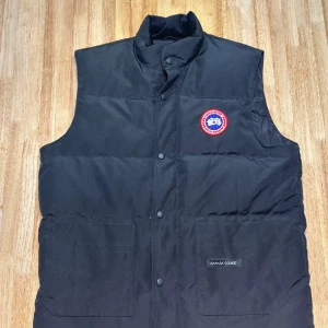 Svart dunväst från Canada Goose - Säljer en svart dunväst från Canada Goose. Västen har knappar och en hög krage för extra värme. Perfekt för kyliga dagar. Finns i XS