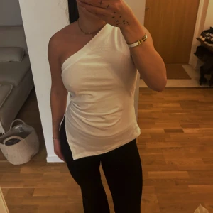 Vit oneshoulder topp - Snygg vit oneshoulder topp med en stilren design. Perfekt för en trendig look. Toppen har en dragkedja på sidan för enkel på- och avtagning.