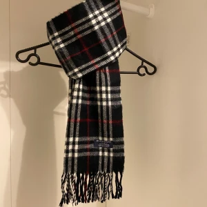 Rutig halsduk från Burberry - Säljer nu denna snygga halsduk från Burberry i svart, Tillverkad i mjuk ull med klassiskt mönster och fransar i ändarna. Perfekt för att hålla dig varm och stilren under kyliga dagar. Hör av er vid minsta funderingar☺️