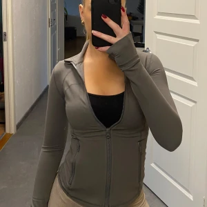 Grå träningskofta - Snygg grå träningskofta med dragkedja och hål till tummen. Finns även en ficka vid armen där man kan ha airpods eller nycklar medans man tränar. Sitter som en smäck och bra material. Oanvänd och prislapp kvar