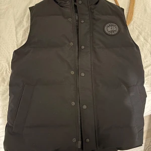 Svart dunväst från Canada Goose - Säljer en svart dunväst från Canada Goose med hög krage och knappar. Västen har en broderad logotyp på bröstet och praktiska fickor framtill. Perfekt för kyliga dagar. Helt oanvänd , pris kan diskuteras👍