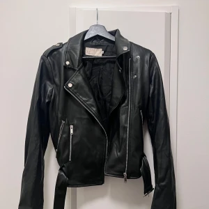 Svart skinnjacka med dragkedja - Snygg svart skinnjacka från Nelly Jackan har dragkedja och knappar samt långa ärmar. 