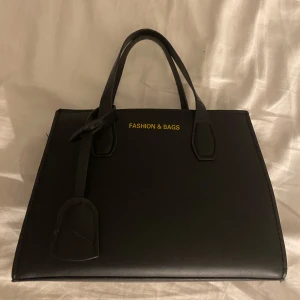 Svart handväska från Fashion & Bags - Elegant svart handväska med stilrena handtag och en praktisk design. Väskan har en diskret logotyp i guld och en dragkedja för säker förvaring. Perfekt för att bära dina dagliga nödvändigheter med stil. Aldrig använd. 