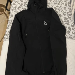 Svart vindjacka från Haglöfs - Säljer en svart jacka från Haglöfs med dragkedja och huva. Perfekt för utomhusaktiviteter med sin stilrena design och funktionella detaljer. Jackan har långa ärmar och är tillverkad i ett slitstarkt material.