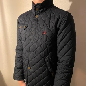 Svart quiltad jacka från Polo Ralph Lauren - Säljer en stilren svart quiltad jacka från Polo Ralph Lauren. Jackan har en klassisk design med knappar och en liten broderad logga på bröstet. Tyvärr är 3 knappar borta men den funkar perfekt att stänga ändå! Om ni vill ha mer information skriv till oss!