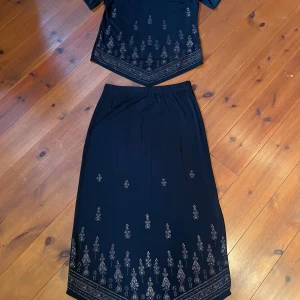 Svart kjol och top med mönster - Elegant svart kjol och top med subtilt mönster i nederkant. Toppen har korta ärmar och en klassisk passform som passar perfekt för både formella och avslappnade tillfällen.