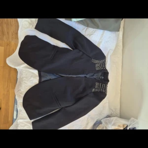 Svart kavaj med nitar - Snygg svart kavaj med coola nitar på kragen. Perfekt för att ge din outfit en edgy touch. Kavajen har en klassisk passform och långa ärmar.