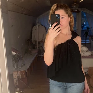 Svart axelbandslös topp - Snygg svart axelbandslös topp med volangdetaljer. Styla den exakt som du vill. Toppen har en luftig passform och är tillverkad i ett mjukt material.