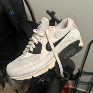 Vita Nike Air Max sneakers - Säljer ett par vita Nike Air Max sneakers med svart swoosh och synlig luftdämpning i sulan. Skorna har snörning och en stilren design som passar perfekt för vardagsbruk. 