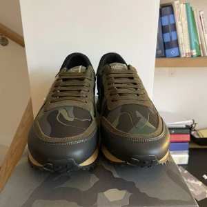 Valentino Rockrunner  - Snygga Valentino Rockrunner sneakers i ett coolt camouflage-mönster. Skorna har en kombination av gröna och svarta nyanser med en beige sula. De är tillverkade i en mix av skinn och syntetmaterial och har klassisk snörning. Perfekta för en trendig look. öppen för bud