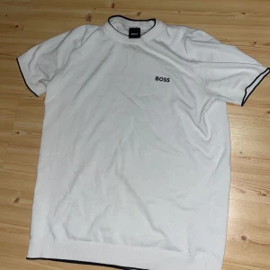 Vit t-shirt från Boss - Snygg vit t-shirt från Boss med svart kantdetalj vid ärmar och hals. Den har en enkel och stilren design med korta ärmar och Boss-loggan broderad på bröstet. Perfekt för en avslappnad och stilren look.