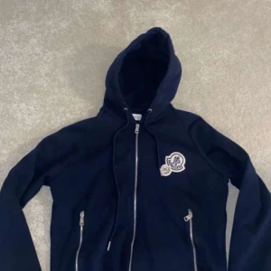 Svart huvtröja från Moncler - Säljer en svart huvtröja från Moncler med dragkedja och två fickor framtill. Tröjan har en broderad logga på bröstet och justerbar huva. 