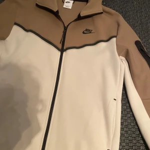 Beige och vit jacka från Nike - Säljer en snygg beige och vit jacka från Nike med dragkedja framtill och en stilren design. Jackan har långa ärmar och en diskret Nike-logga på bröstet. Perfekt för en sportig look!