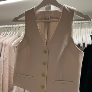 Beige väst från H&M - Snygg väst från H&M med knappar framtill. Perfekt för att lägga till en stilren touch till din outfit. Västen har en klassisk design med två fickor och en v-ringad halslinje. Endast använd en gång 🤎🤎