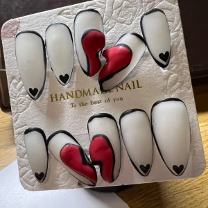 Handgjorda lösnaglar med hjärtdetaljer - Snygga handgjorda lösnaglar i vitt med svarta kanter och små svarta hjärtan. Två naglar har en röd hjärtdesign i mitten. Perfekt för en unik och stilren look.