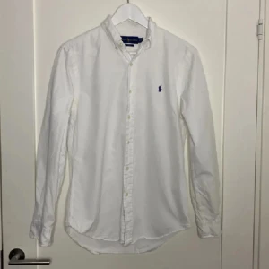 Ralph lauren skjorta  - Säljer en klassisk vit skjorta från Ralph Lauren med broderad logga på bröstet. Skjortan har långa ärmar och knappar framtill. Perfekt för en stilren look. Längd 60 cm