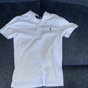 Vit kortärmad t-shirt från Ralph Lauren - Säljer en stilren vit kortärmad t-shirt från Ralph Lauren med en klassisk krage och knappdetalj. Den har en broderad logotyp på bröstet och är i slim fit. Perfekt för en avslappnad och snygg look. Den är köpt för 1000kr. Barnstorlek L. Kommer passa dig som är runt 170