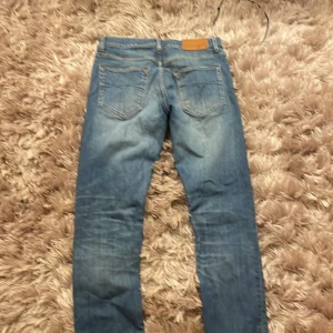 Blå jeans från Tiger of Sweden - Snygga blå jeans från Tiger of Sweden med klassisk femficksdesign och knappgylf. Tillverkade i Italien, dessa jeans har en stilren look med lätt slitna detaljer för en trendig touch.