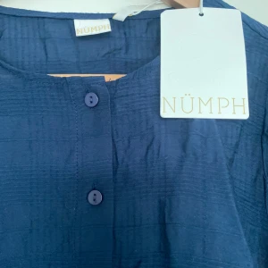 Mörkblå blus från Nümph - Säljer en stilren mörkblå blus från Nümph med knappar framtill. Blusen har en rund halsringning och långa ärmar, perfekt för en chic och avslappnad look. Materialet har en subtil textur som ger extra karaktär.
