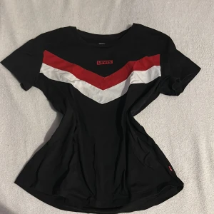 Svart t-shirt från Levi's - Snygg svart t-shirt från Levi's med ett coolt rött och vitt V-format mönster på bröstet. Perfekt för en avslappnad stil. Tillverkad i mjukt material för extra komfort. Säljer även likadan fast i vit🫶