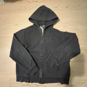 Svart zip hoodie från Polo Ralph Lauren - Säljer en svart hoodie från Polo Ralph Lauren med dragkedja och huva. Den har långa ärmar och en mysig insida. Perfekt för en avslappnad stil.