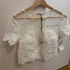 Vit spetsblus från Ida Sjöstedt - Blusen har korta ärmar och ett vackert prickigt mönster på övre delen. Perfekt för en stilren look med fina spetsdetaljer.