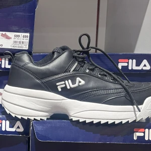 Svarta sneakers från Fila - Snygga svarta sneakers från Fila med vit sula och snörning. Perfekta för en sportig look med en bekväm passform. Fila-loggan syns tydligt på sidan av skorna.